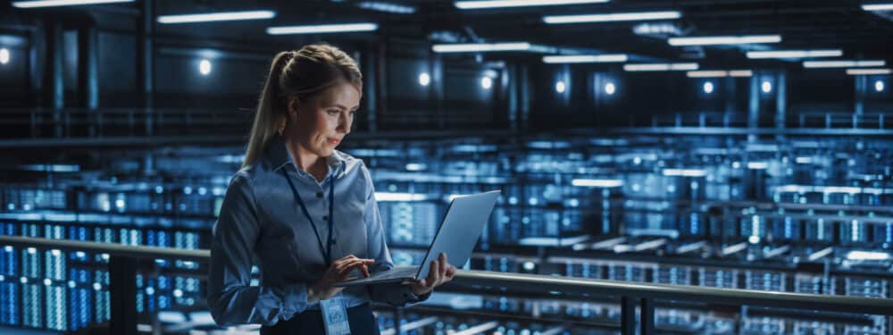 Professional woman in business attire poses in front of a server room with rows of servers