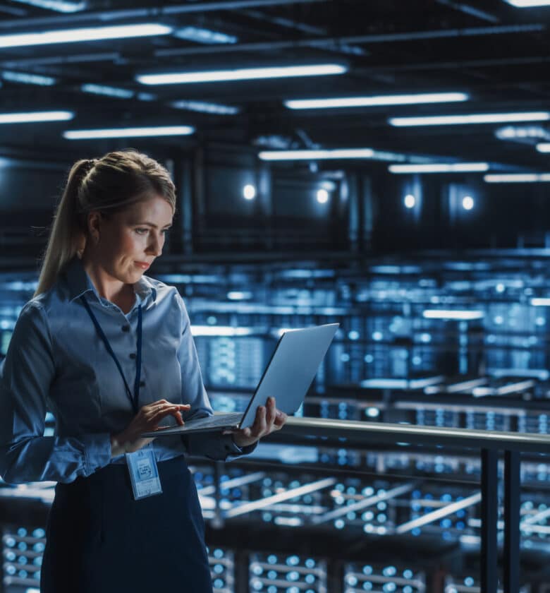 Professional woman in business attire poses in front of a server room with rows of servers