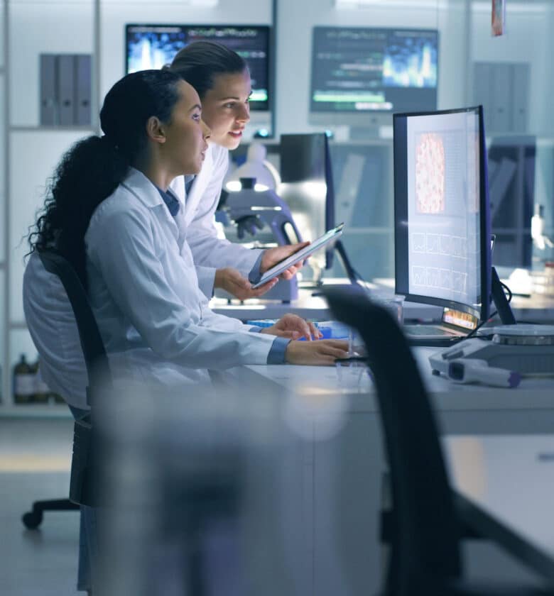 Two female scientists in lab coats examining information on computer screens