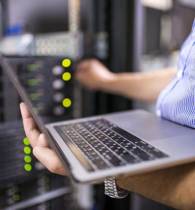 Person holding laptop in front of server rack, checking network connections